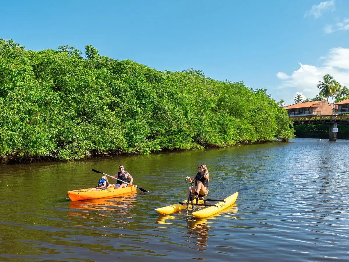 Família de caiaque e hóspede de pedalinho no Rio Maragogi, com o mangue na margem e um bloco do resort ao fundo