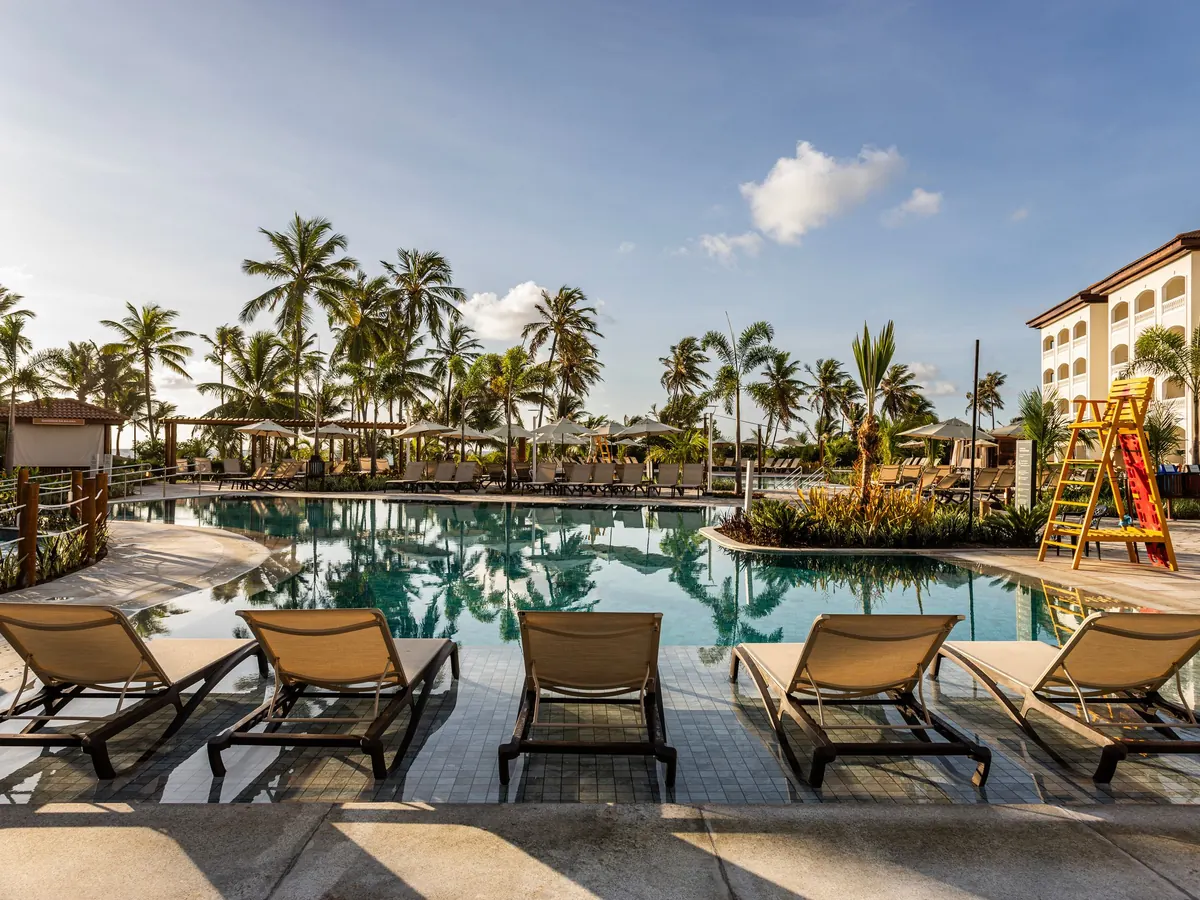 Espreguiçadeiras na parte rasa da piscina do Sauípe Sol Grand Premium, com coqueiros e o prédio do hotel ao lado