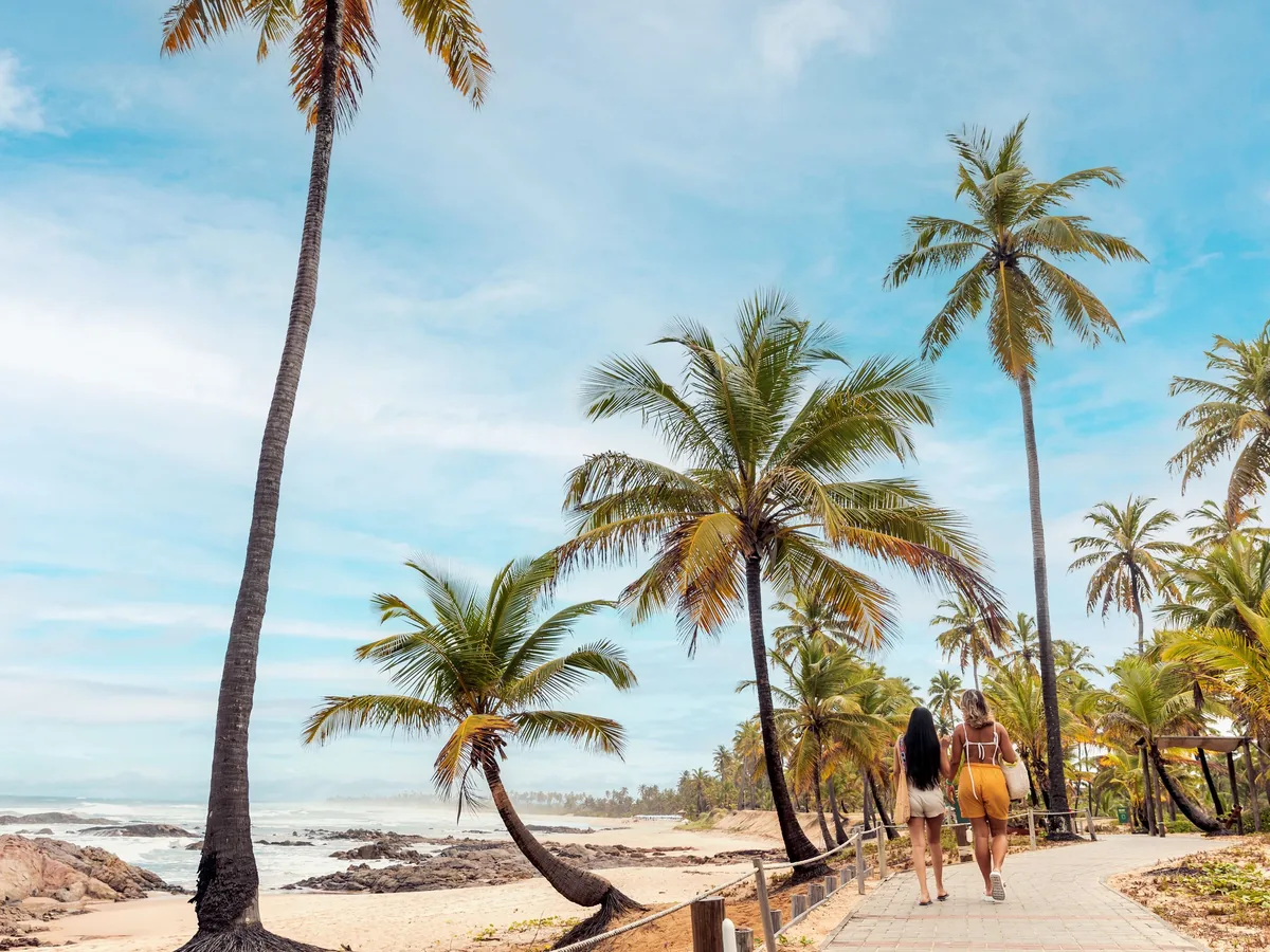 Duas mulheres caminhando no calçadão entre os coqueiros e a praia da Costa do Sauípe