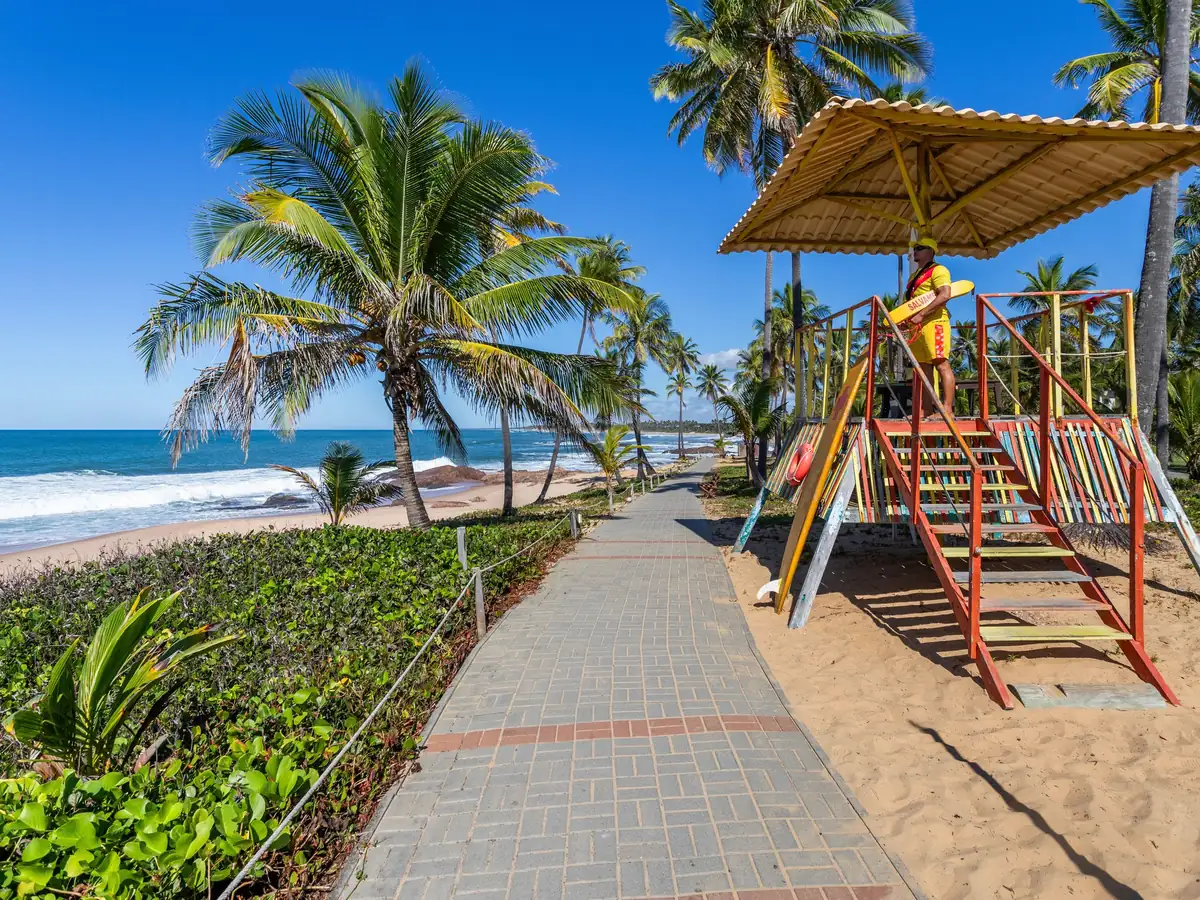 Calçadão da orla da Costa do Sauípe entre a praia e os coqueiros, com um salva-vidas no posto