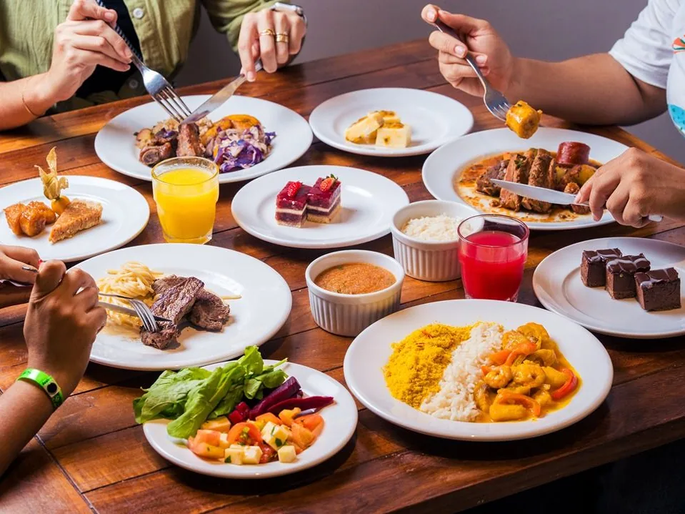 Hóspedes almoçando numa mesa com moqueca de camarão, carnes, salada e sobremesas