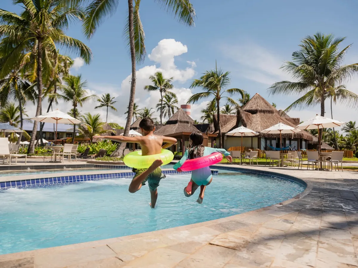 Crianças pulando com boias na piscina do Summerville, com bangalôs de palha e coqueiros ao fundo