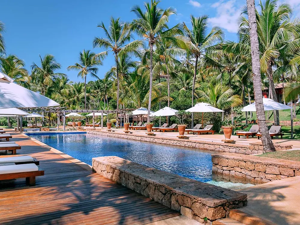 Piscina do Txai com deque de madeira, espreguiçadeiras e guarda-sóis brancos entre coqueiros