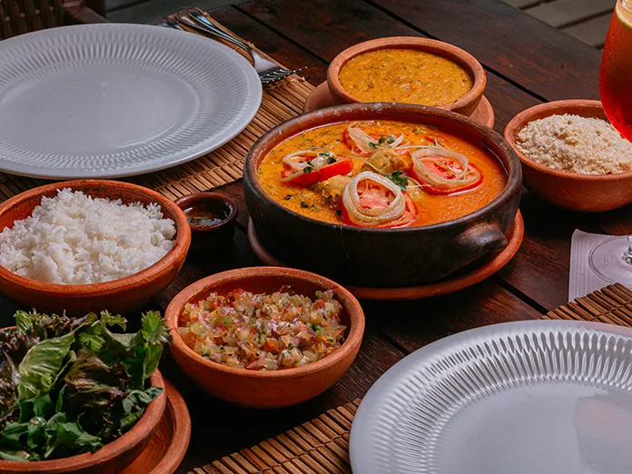 Moqueca na panela de barro com arroz, farofa, salada e outros acompanhamentos numa mesa do Txai