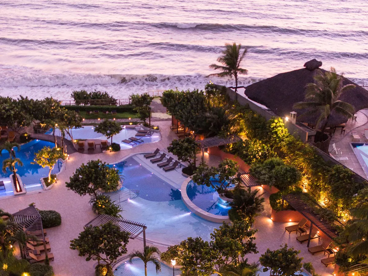 O Vogal Luxury Beach Hotel visto do alto ao entardecer, com as piscinas iluminadas e o mar logo atrás