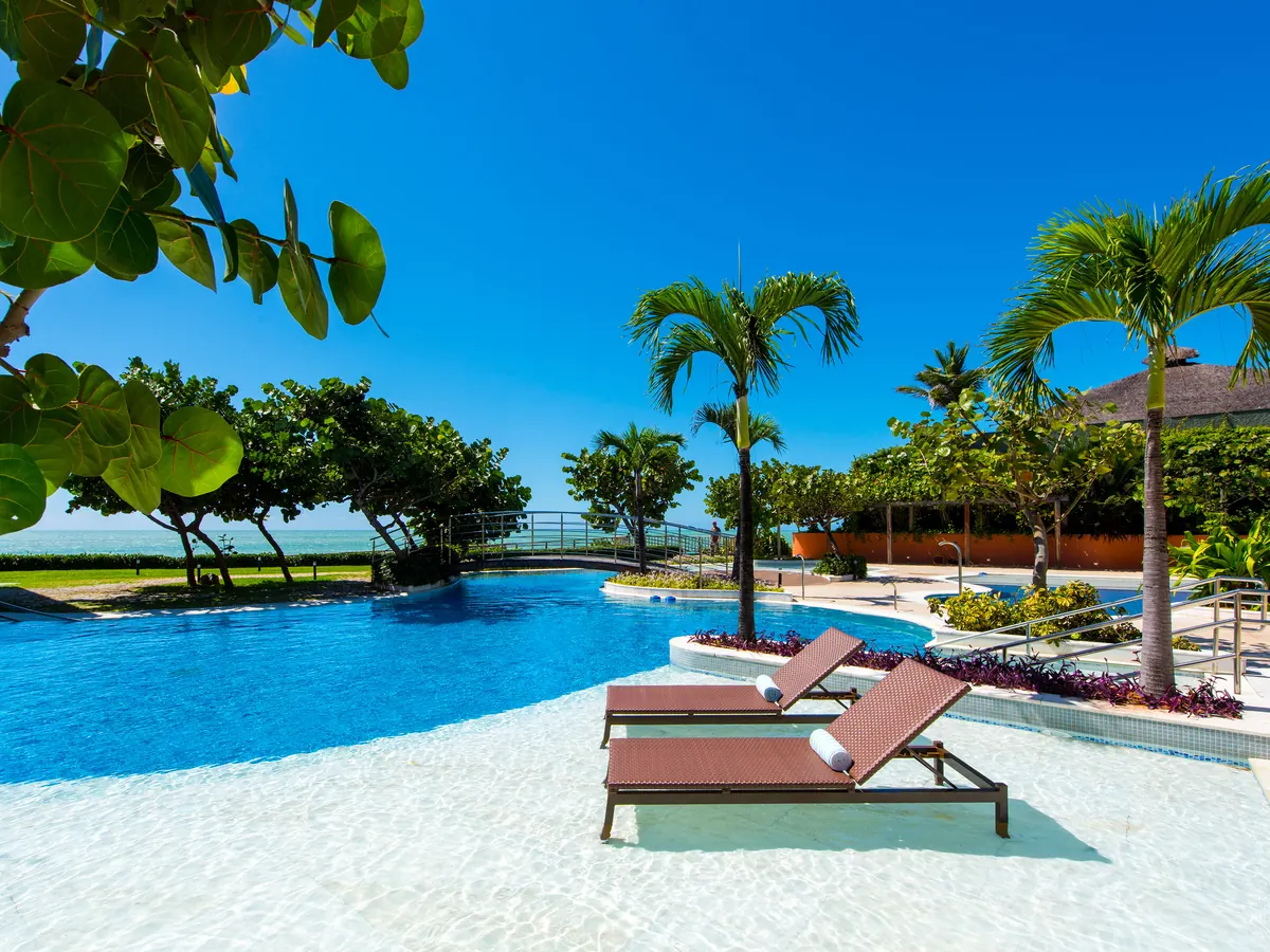 Piscina do Vogal Luxury Beach Hotel com espreguiçadeiras dentro da água rasa, coqueiros e uma ponte ao fundo